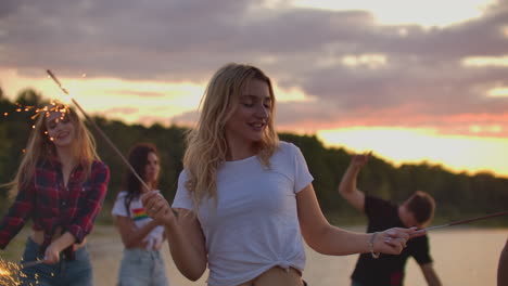 The-blonde-girl-with-nude-waist-is-dancing-with-big-bengal-lights-in-her-hands-on-the-sand-beach-with-her-friends.-This-is-wonderful-summer-evening-on-the-open-air-party.